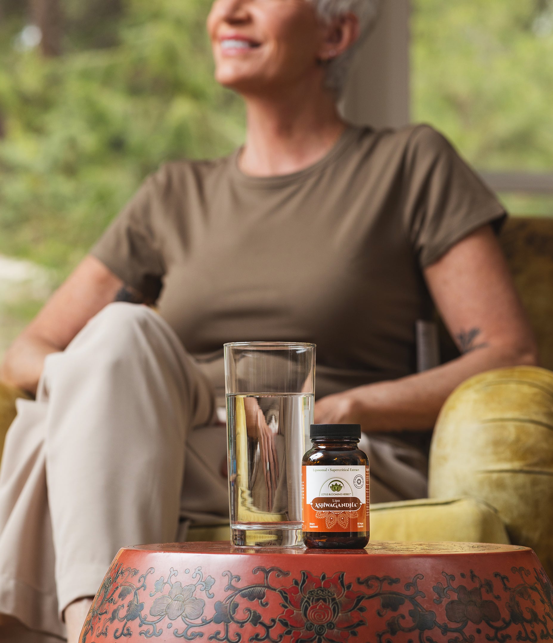 Woman sitting on a chair with a glass of water and ashwagandha supplement bottle on a table.
