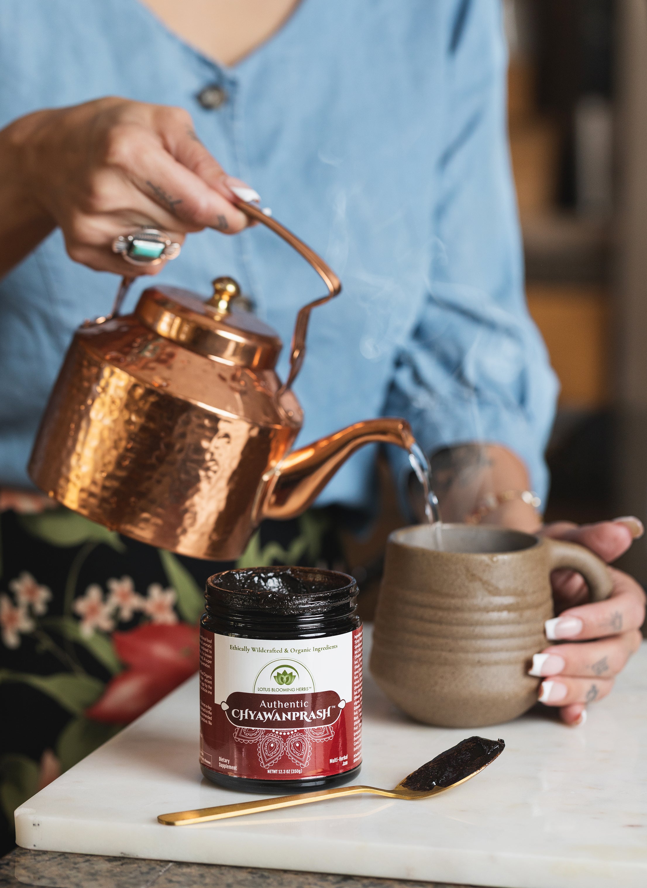 Person pouring tea from a copper kettle into a mug with a jar of chyawanprash on a marble surface.